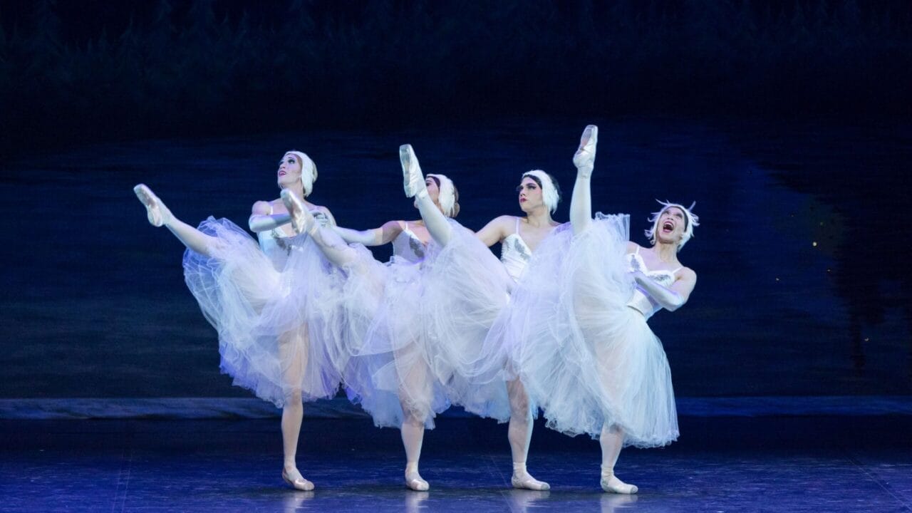 Four ballet dancers in white tutus and headpieces from Les Ballets Trockadero perform on stage, each lifting one leg high in perfect coordination, set against a dark, forested backdrop reminiscent of Monte Carlo’s elegant ballet tradition.