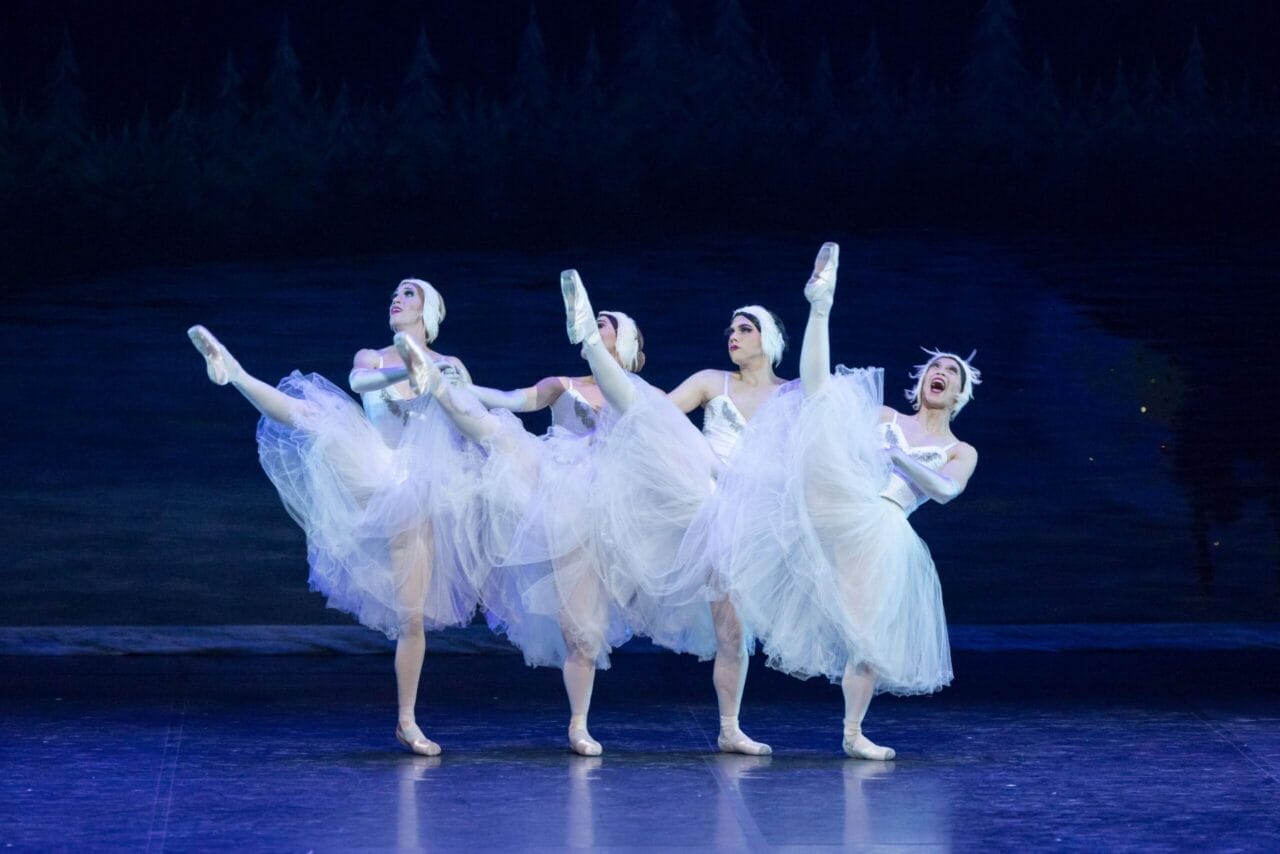 Four ballet dancers in white tutus and headpieces from Les Ballets Trockadero perform on stage, each lifting one leg high in perfect coordination, set against a dark, forested backdrop reminiscent of Monte Carlo’s elegant ballet tradition.