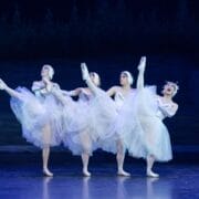 Four ballet dancers in white tutus and headpieces from Les Ballets Trockadero perform on stage, each lifting one leg high in perfect coordination, set against a dark, forested backdrop reminiscent of Monte Carlo’s elegant ballet tradition.