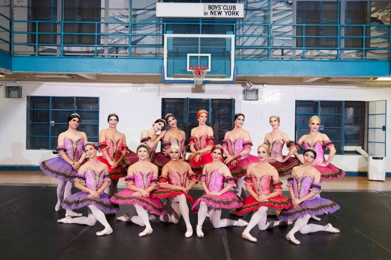 A group of ballet dancers in colorful tutus poses together on a basketball court in a gymnasium, channeling Les Ballets Trockadero de Monte Carlo, with a “Boys Club of New York” sign above the hoop.