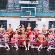 A group of ballet dancers in colorful tutus poses together on a basketball court in a gymnasium, channeling Les Ballets Trockadero de Monte Carlo, with a “Boys Club of New York” sign above the hoop.