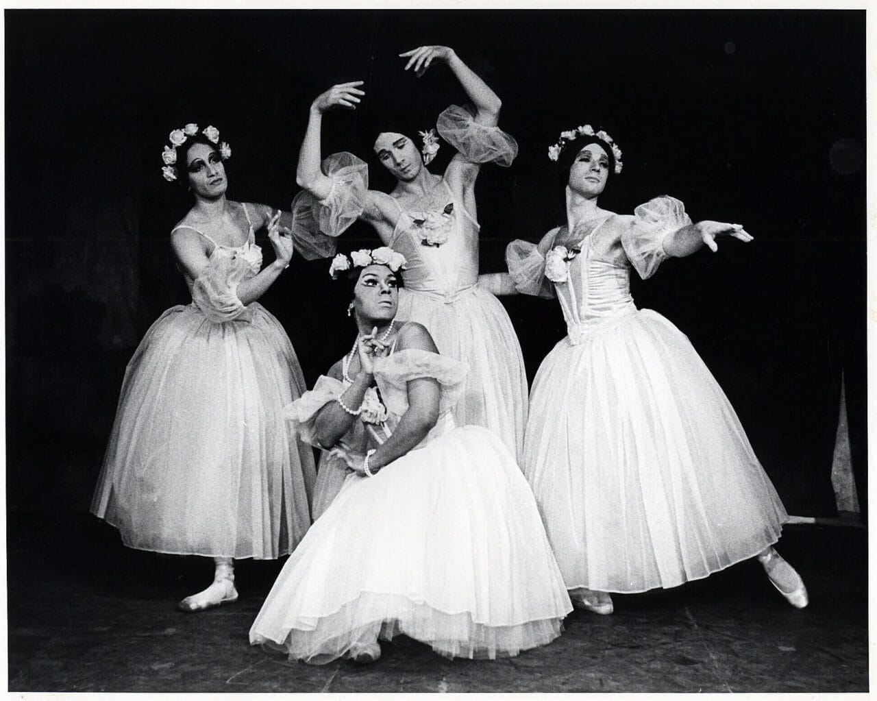 Four ballet dancers in white tutus and floral headpieces pose on stage against a dark backdrop, their expressive arms capturing the playful spirit of Les Ballets Trockadero de Monte Carlo.