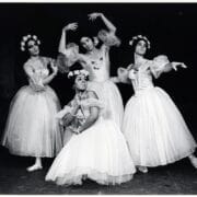 Four ballet dancers in white tutus and floral headpieces pose on stage against a dark backdrop, their expressive arms capturing the playful spirit of Les Ballets Trockadero de Monte Carlo.