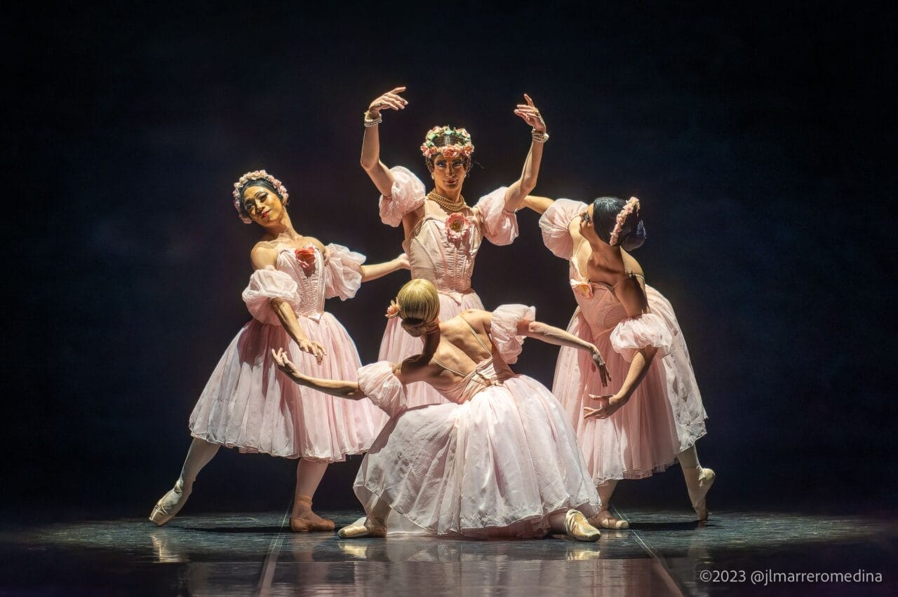 Four ballet dancers in pink tutus and floral headpieces, inspired by Les Ballets Trockadero, pose gracefully on stage under dramatic lighting, capturing the elegance of ballet with a hint of Monte Carlo flair.