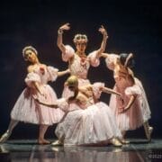 Four ballet dancers in pink tutus and floral headpieces, inspired by Les Ballets Trockadero, pose gracefully on stage under dramatic lighting, capturing the elegance of ballet with a hint of Monte Carlo flair.