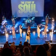 A band and singers perform on stage with "The Story of Soul" displayed behind them, as sparklers light up each side and the audience enjoys the soulful story from their seats.