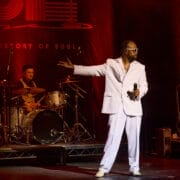 A singer in a white suit and sunglasses performs on stage with a drummer and guitarist, holding a microphone and gesturing with his hand under red lighting, delivering a soulful story through his powerful vocals.