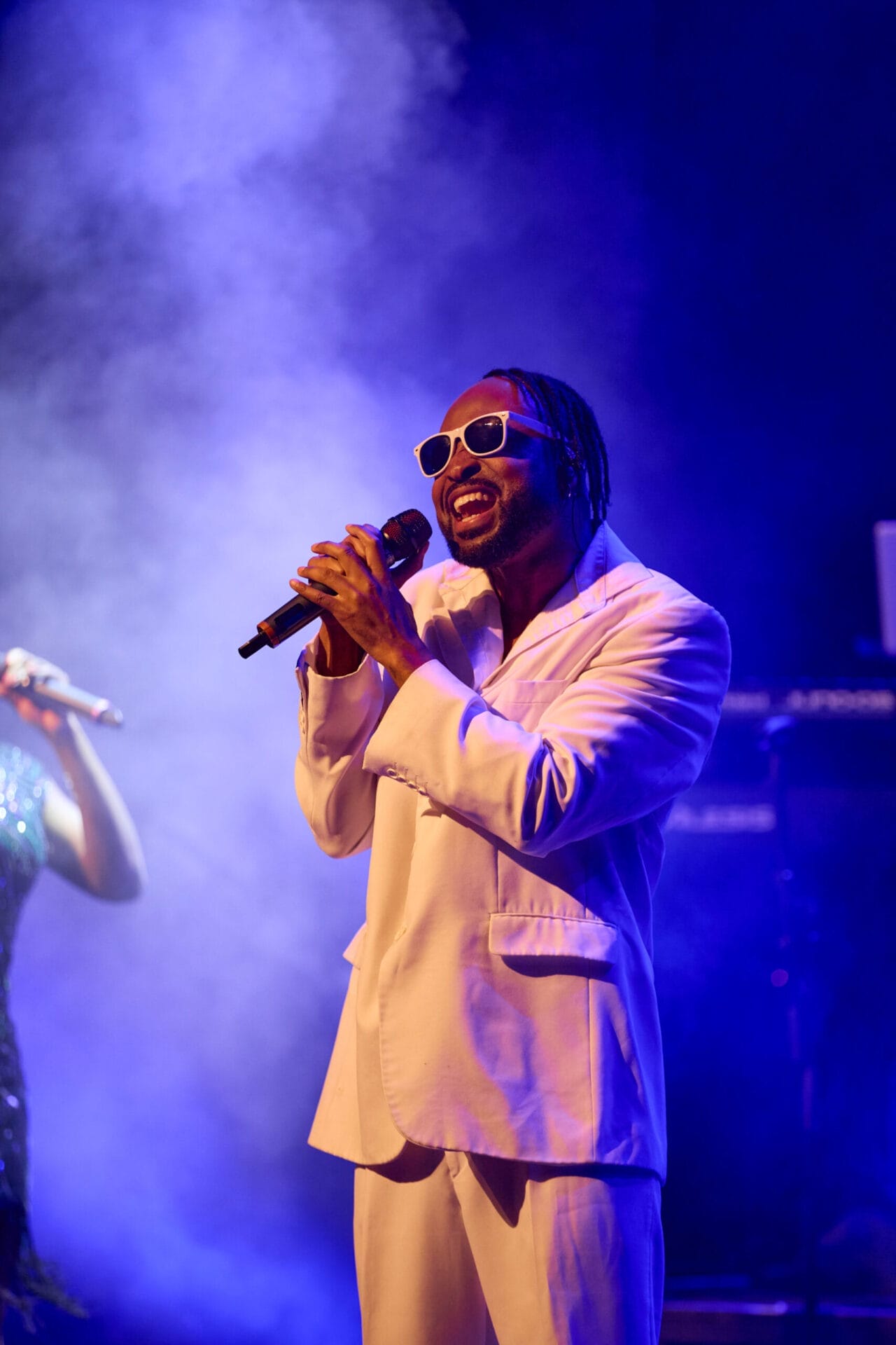 A man wearing a white suit and sunglasses sings into a microphone on stage, surrounded by blue and purple lighting and smoke effects—bringing the Story of Soul to life with every powerful note.
