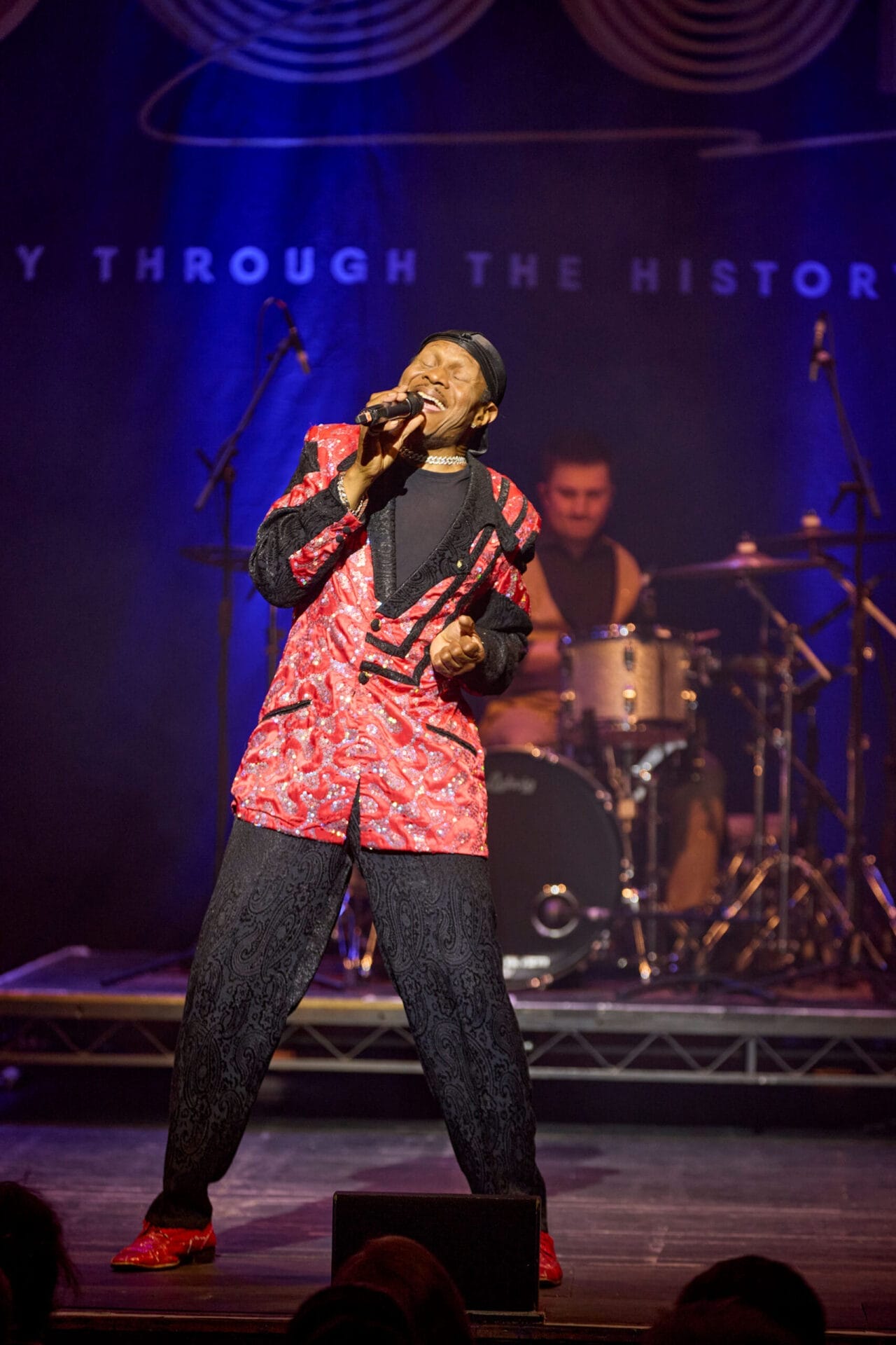 A singer in a red and black patterned suit performs a soulful set on stage, sharing the story of soul, with a drummer in the background under blue stage lighting.