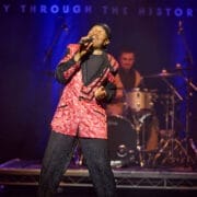 A singer in a red and black patterned suit performs a soulful set on stage, sharing the story of soul, with a drummer in the background under blue stage lighting.