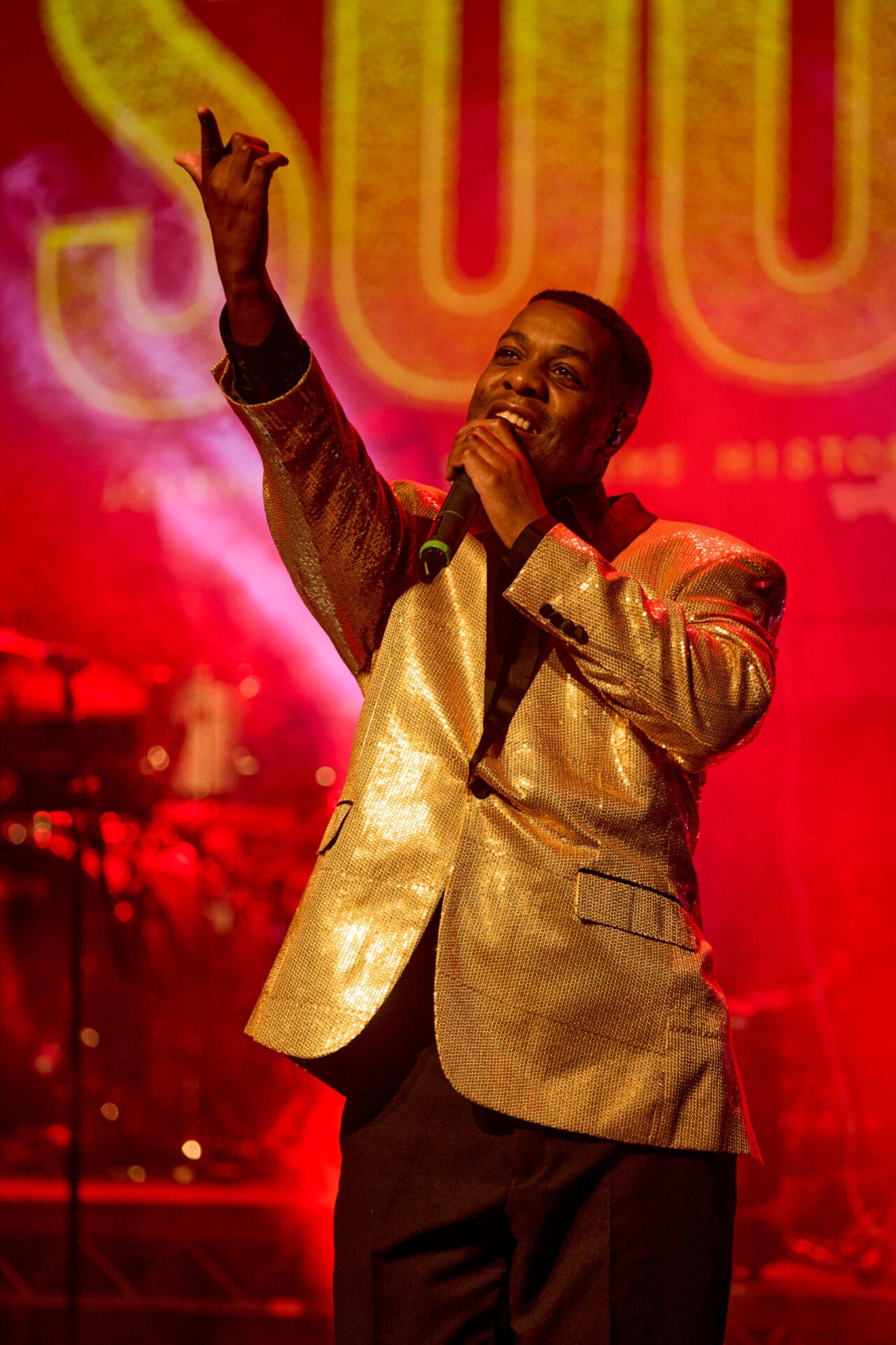 A singer in a shiny gold jacket performs a soul story on stage with a microphone, set against a red background with large illuminated letters.