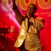 A singer in a shiny gold jacket performs a soul story on stage with a microphone, set against a red background with large illuminated letters.