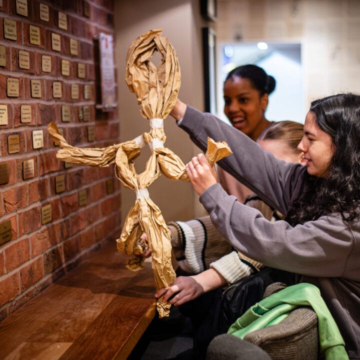 Two young women create a tall figure made of crumpled brown paper in a warmly lit room, reminiscent of creative activities found in educational workshops or schools workshops.