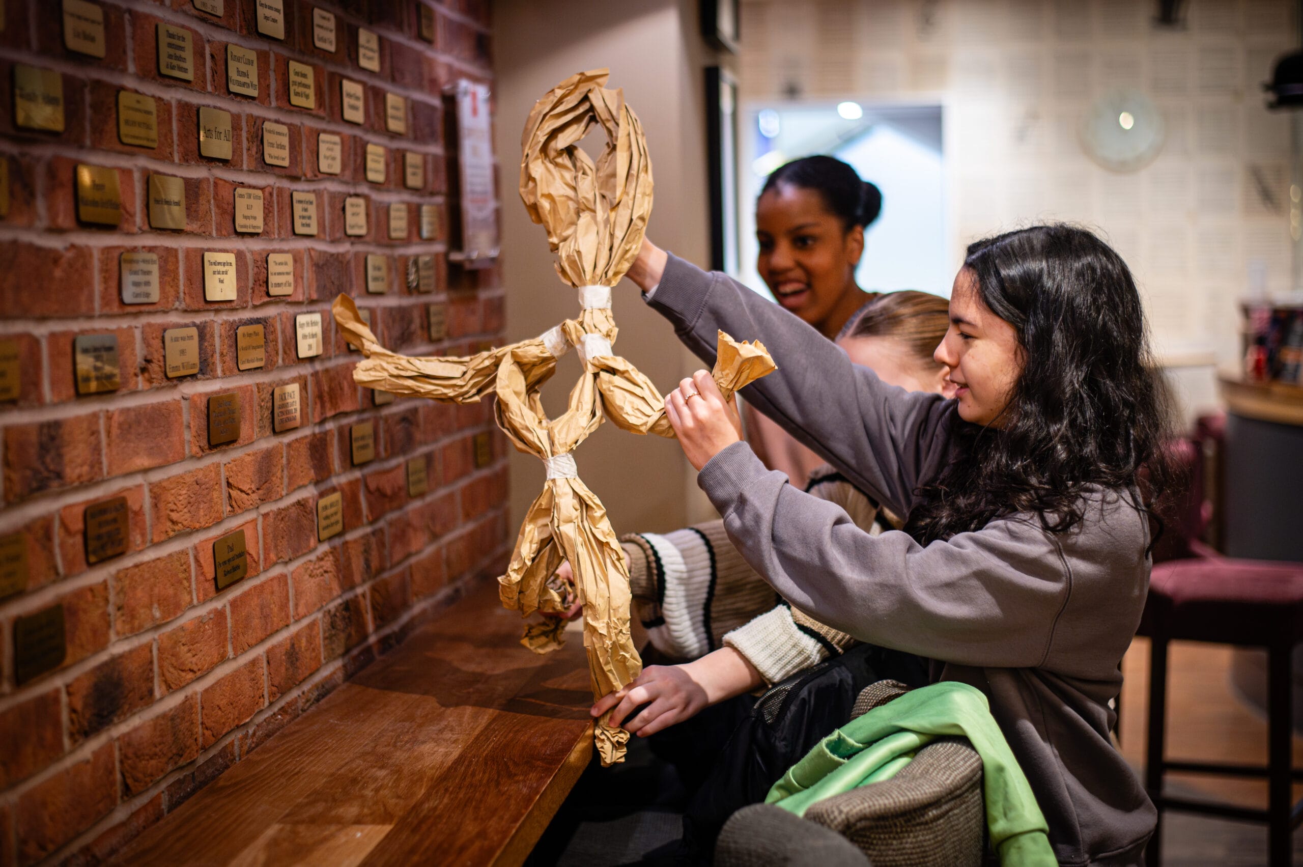 Two young women create a tall figure made of crumpled brown paper in a warmly lit room, reminiscent of creative activities found in educational workshops or schools workshops.