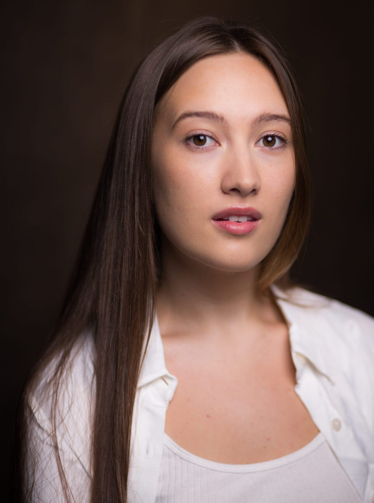 A young woman with straight brown hair and light skin looks at the camera. She is wearing a white shirt over a white top.