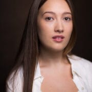 A young woman with straight brown hair and light skin looks at the camera. She is wearing a white shirt over a white top.