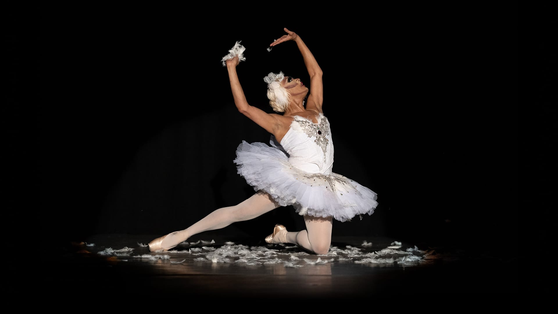 Ballet dancer in a white tutu poses dramatically on one knee on stage, surrounded by scattered white feathers and a dark background, evoking the elegance of Monte Carlo’s renowned ballet scene.