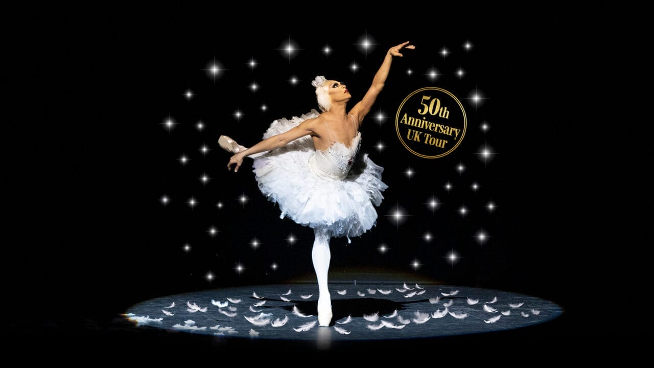 A ballerina in a white tutu poses on stage with feathers on the floor and sparkling lights, promoting Les Ballets Trockadero de Monte Carlo's "50th Anniversary UK Tour" and celebrating the artistry of ballet.