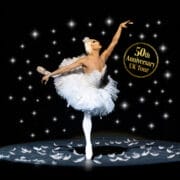 A ballerina in a white tutu poses on stage with feathers on the floor and sparkling lights, promoting Les Ballets Trockadero de Monte Carlo's "50th Anniversary UK Tour" and celebrating the artistry of ballet.