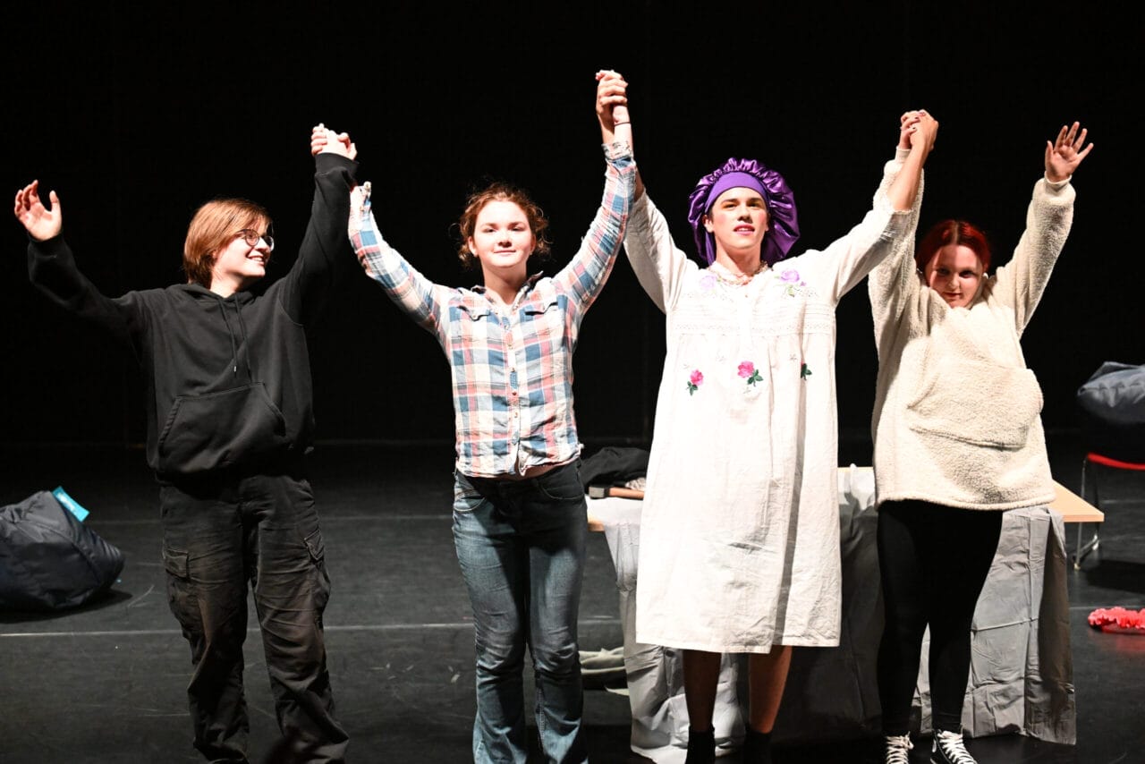 Four people stand on a stage holding hands and raising their arms; one, dressed in a costume, brings Theatre Insights to life, while the others wear casual clothing.