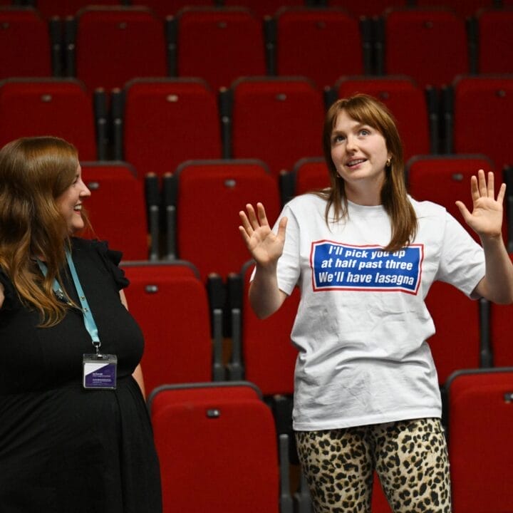 Two women stand and talk in front of empty red theater seats; one gestures with raised hands and wears a shirt with text, while the other looks on and smiles—sharing Theatre Insights in a truly hands-on experience.