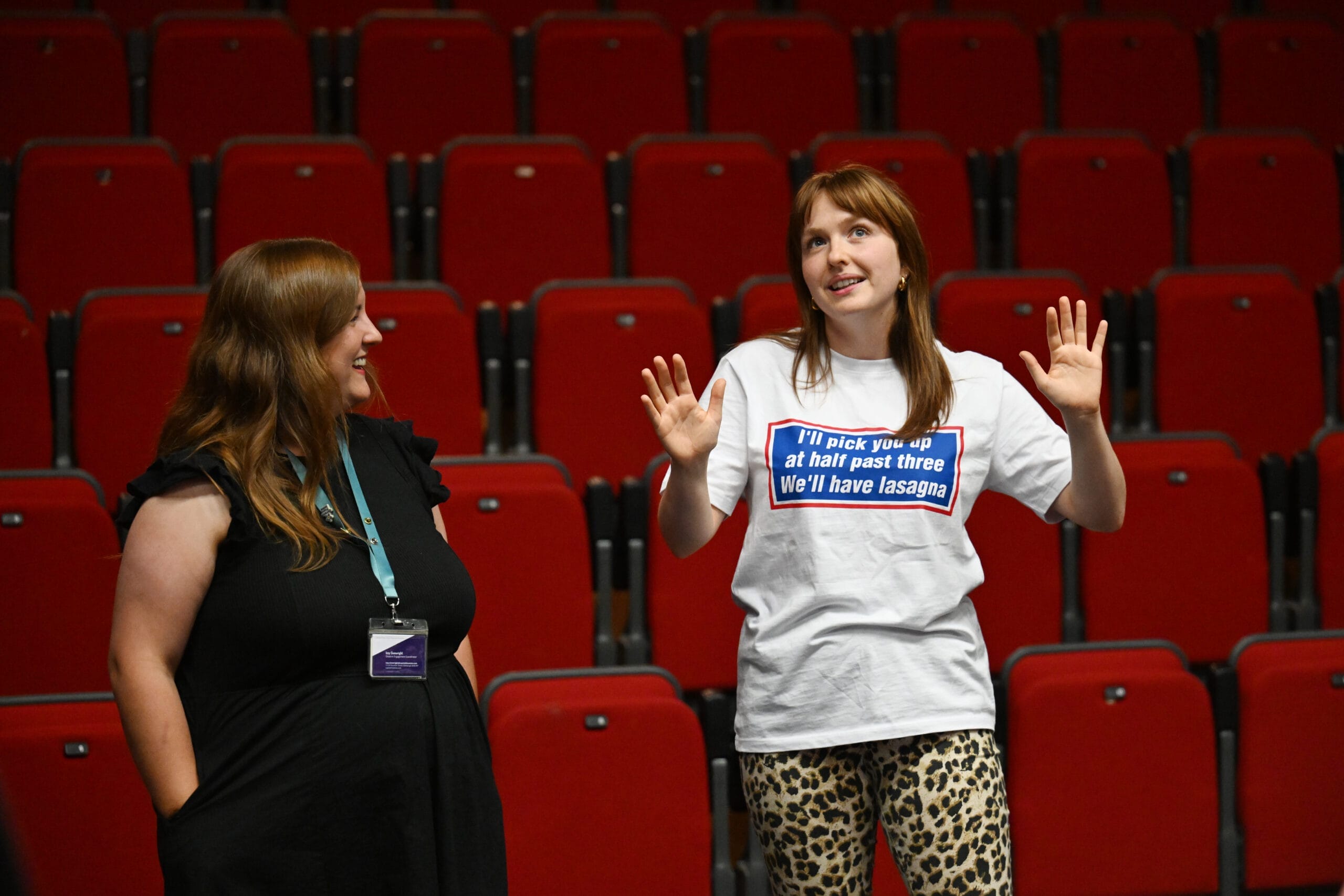 Two women stand and talk in front of empty red theater seats; one gestures with raised hands and wears a shirt with text, while the other looks on and smiles—sharing Theatre Insights in a truly hands-on experience.