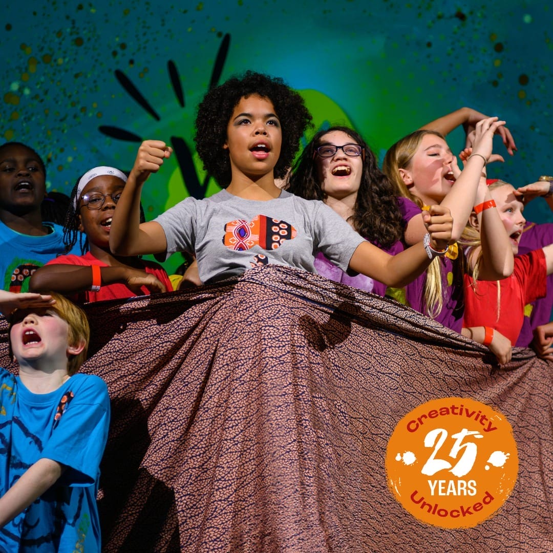 A diverse group of children sing enthusiastically on stage, holding a patterned cloth. A badge reads “Creativity Unlocked: 25 Years,” celebrating the spirit of the Shakespeare Schools Festival.