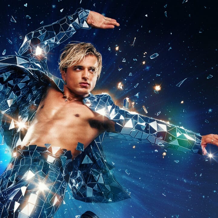 A shirtless man wearing a reflective, mirror-like suit appears to burst through shards of glass against a dark, starry background.