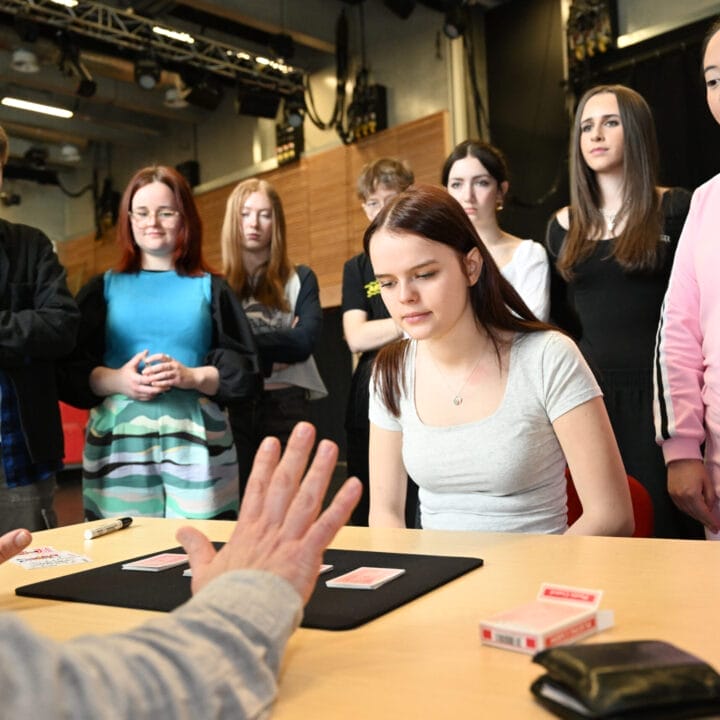 A group of young people watch closely as a person demonstrates a card trick at a table with playing cards.