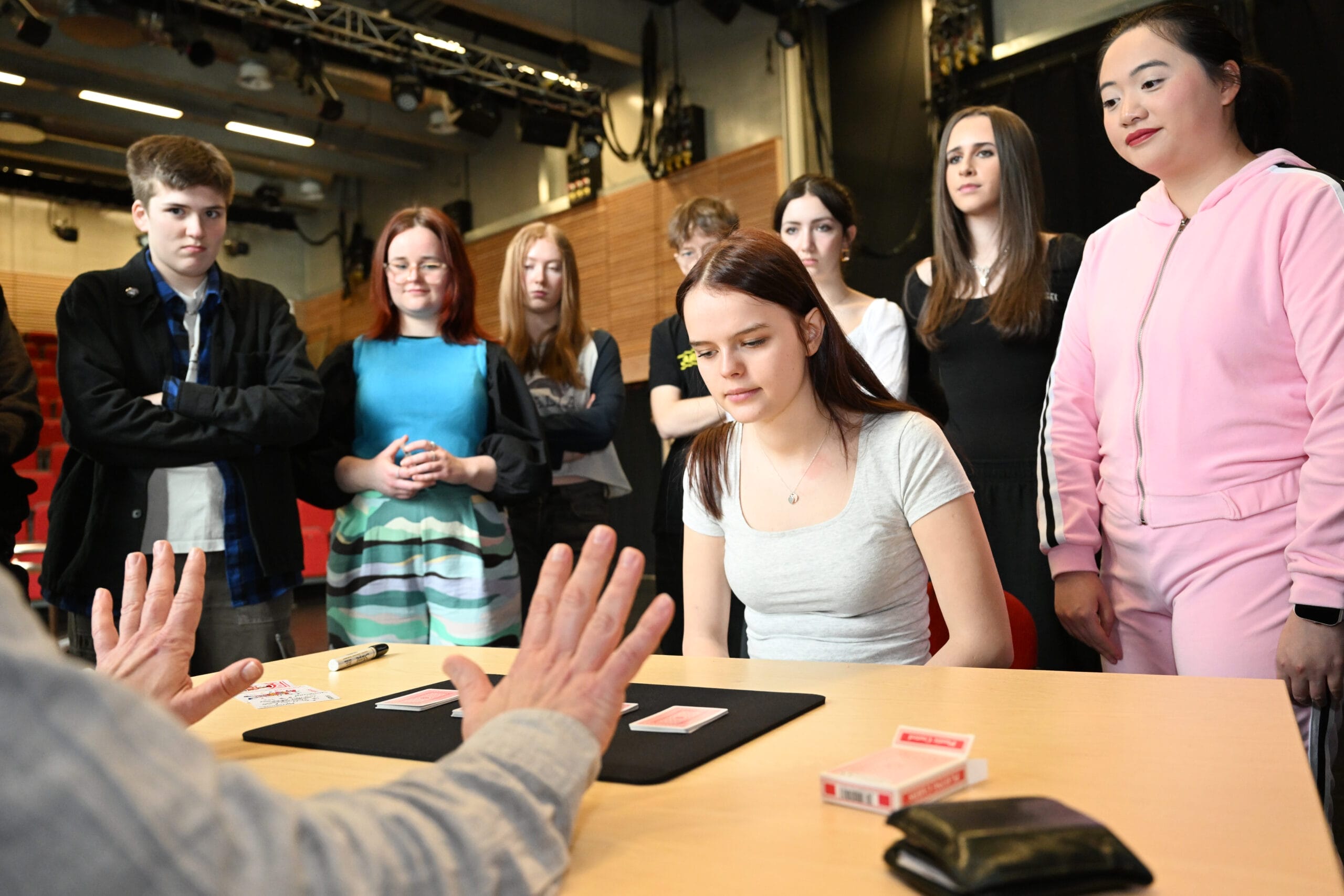 A group of young people watch closely as a person demonstrates a card trick at a table with playing cards.