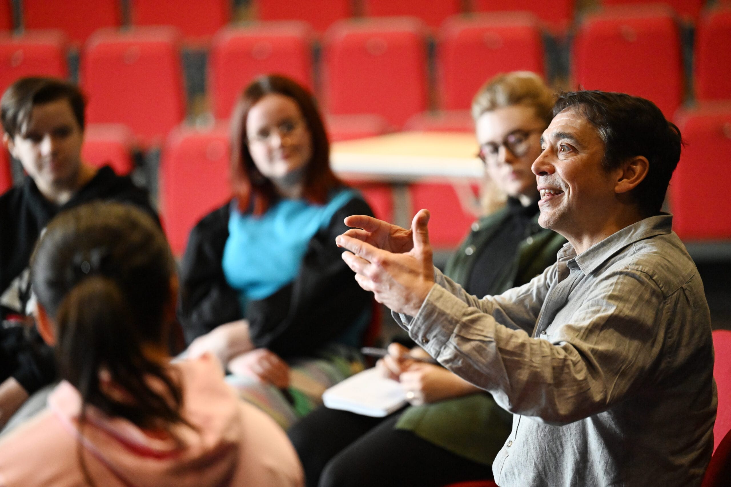 A man speaks to a small group of people seated in a circle in a room with red chairs, as they explore Creative Pathways together while others listen attentively and take notes.