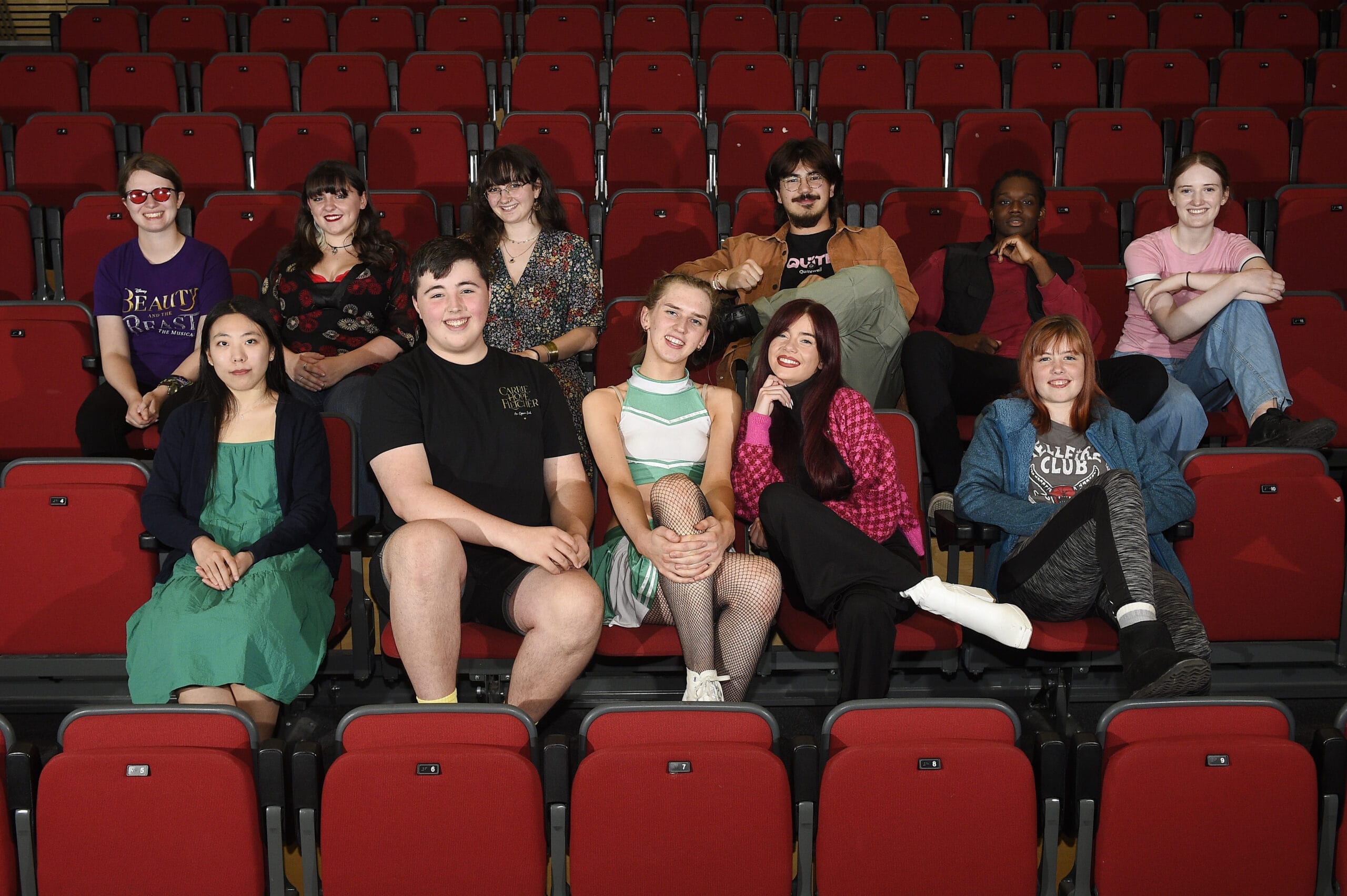 A group of eleven young adults from the Youth Advisory Board sit and pose in red theatre seats, facing the camera and smiling.