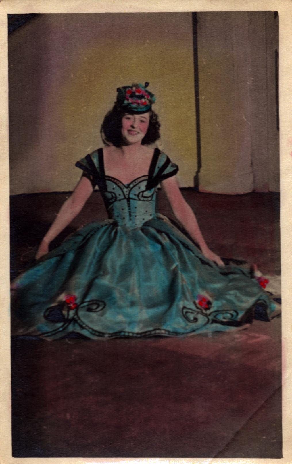 A woman in an off-the-shoulder gown with floral details and a matching headpiece sits on the floor against a plain background. A once black and white photo coloured in blue.