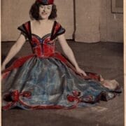 A woman in an off-the-shoulder gown with floral details and a matching headpiece sits on the floor against a plain background. A once black and white photo coloured in red and blue.