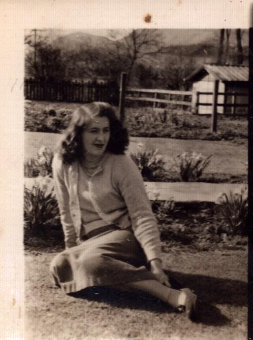 Black and white photo of a woman, Mary Rose, in a cardigan and skirt sits on the grass in a garden with wooden fences and a small shed behind her.
