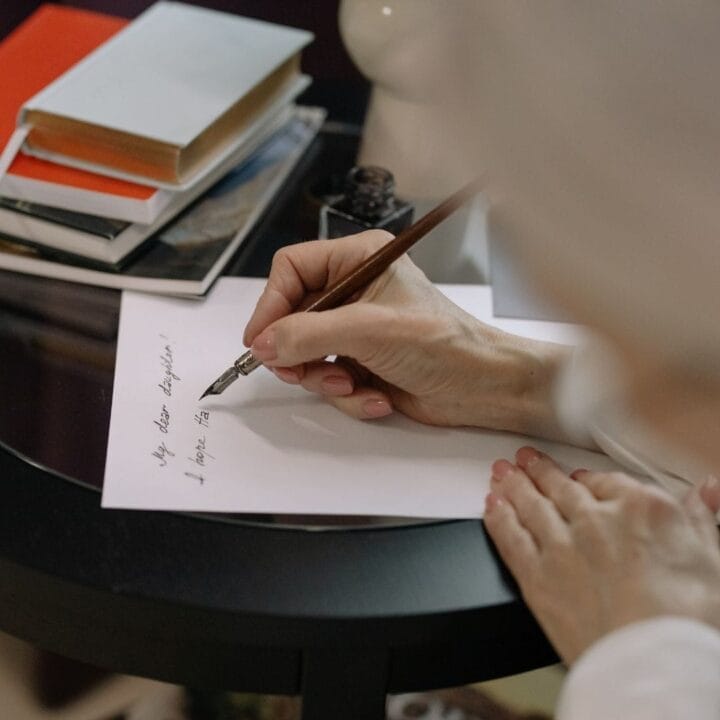 A person writes a note with a pen on white paper at a round black table, stacked notebooks and books in the background—perfect for crafting Relaxing Rhymes.