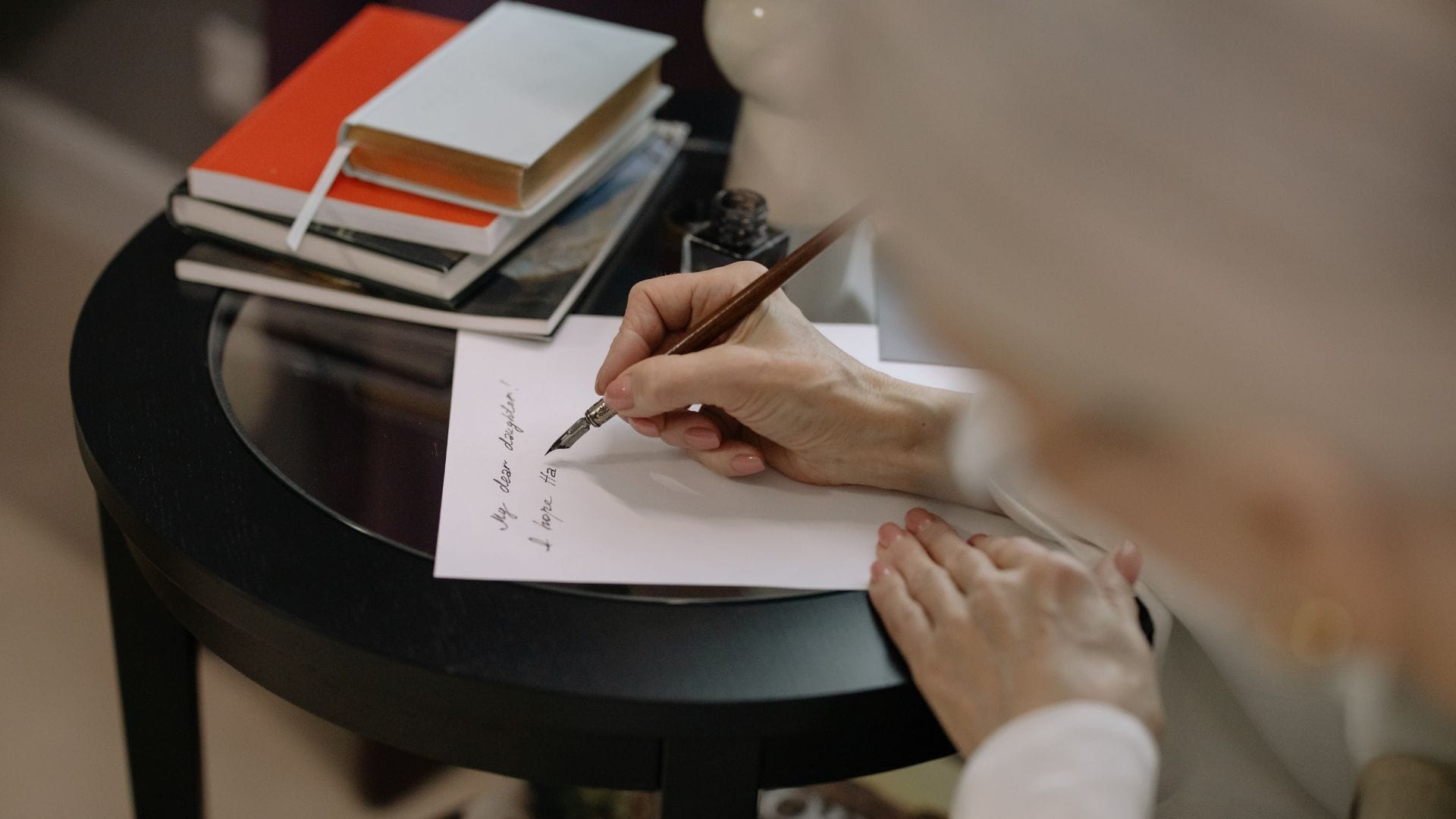 A person writes a note with a pen on white paper at a round black table, stacked notebooks and books in the background—perfect for crafting Relaxing Rhymes.