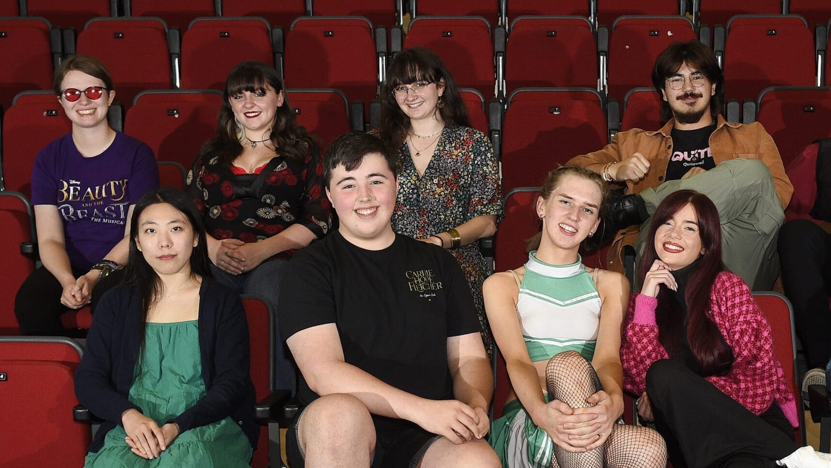 A group of eight young adults sit and pose for a photo in red theater seats, smiling and facing the camera.