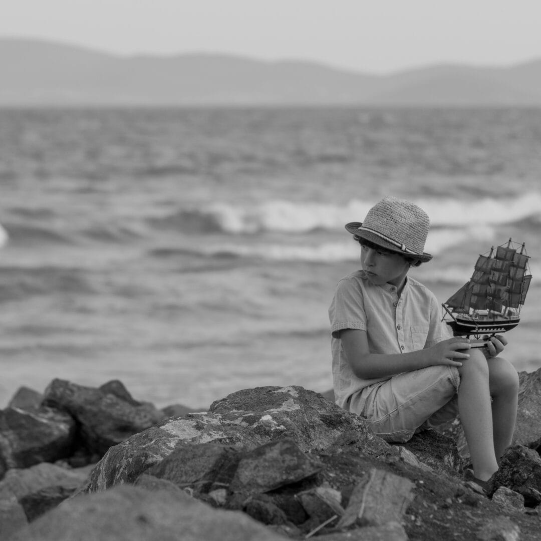 A boy in a hat sits on rocky shore, holding a model ship, looking toward the sea; waves and distant hills are visible in the background. The image is in black and white.
