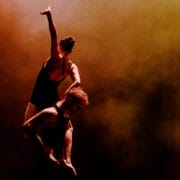 Two dancers perform a balancing act; one stands upright on the other's shoulders with an arm raised, both lit by dramatic orange and yellow stage lighting.