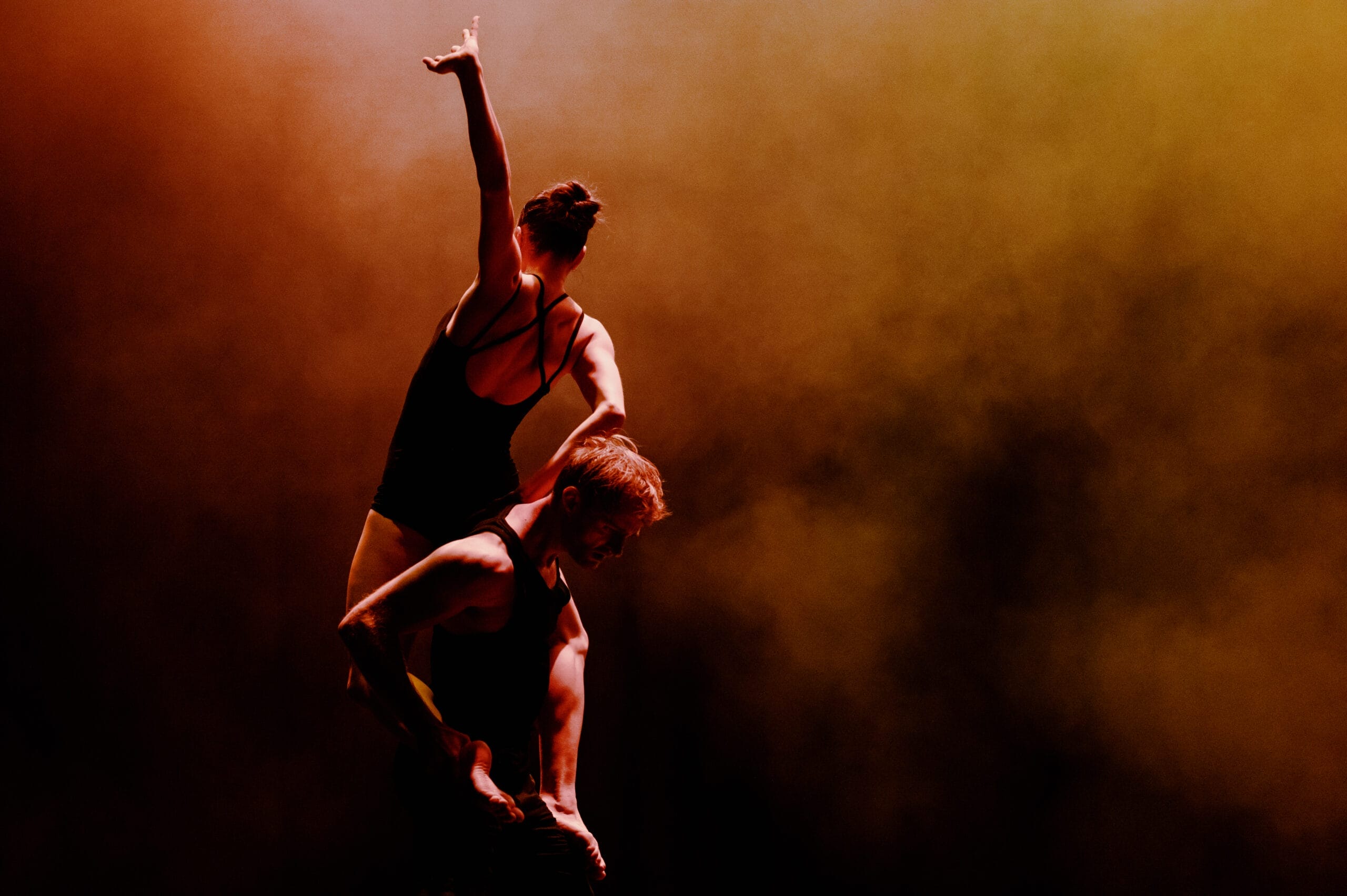 Two dancers perform a balancing act; one stands upright on the other's shoulders with an arm raised, both lit by dramatic orange and yellow stage lighting.