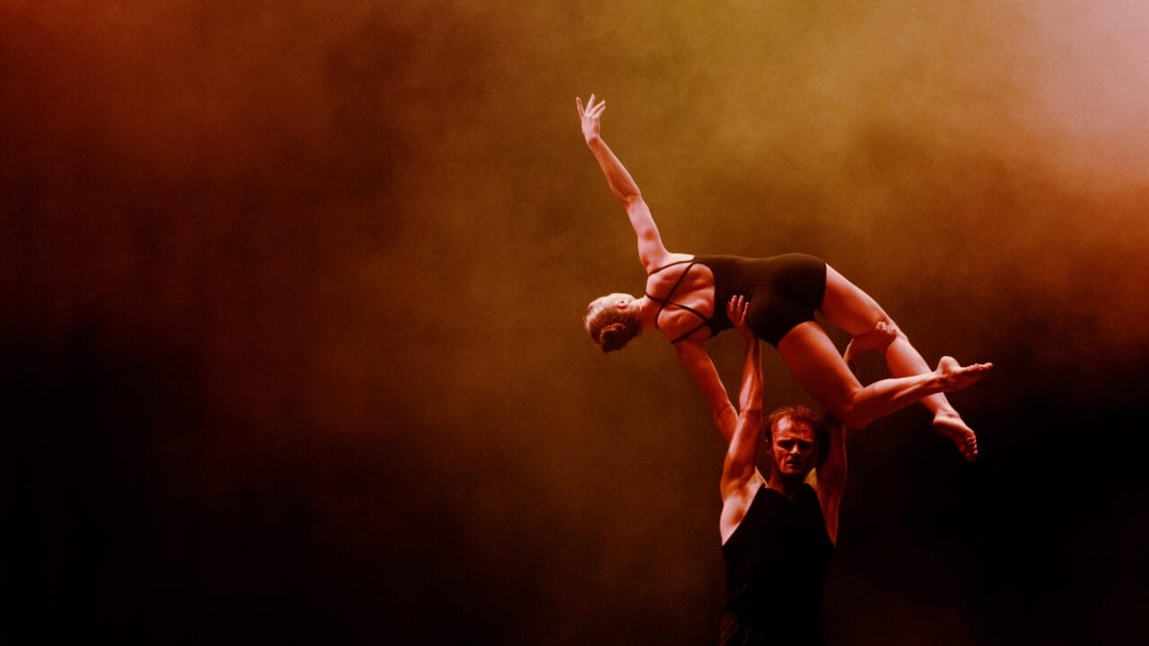 Two acrobats perform a lift on stage, with one person holding another above their head against a smoky, orange-lit background.