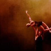 Two acrobats perform a lift on stage, with one person holding another above their head against a smoky, orange-lit background.