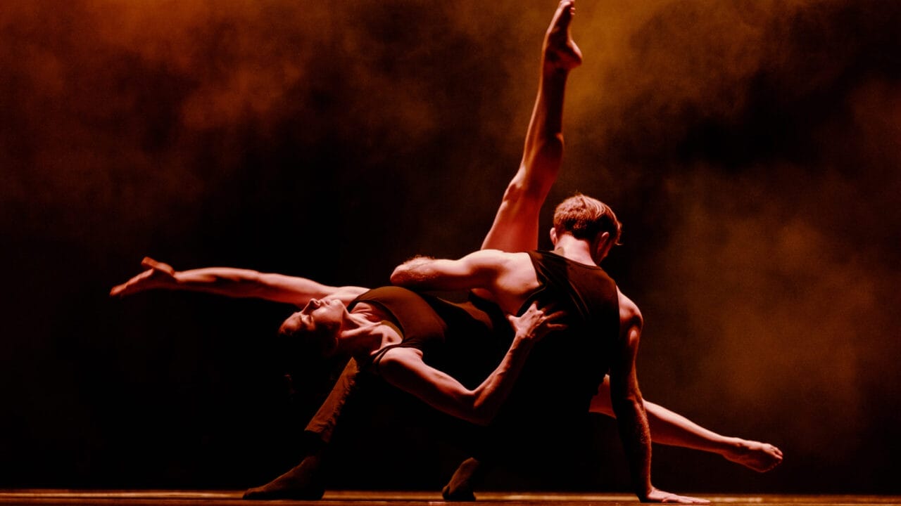 Two dancers perform a dramatic pose on stage, one arching backward with an extended leg, against a dark background with orange lighting and smoke effects.