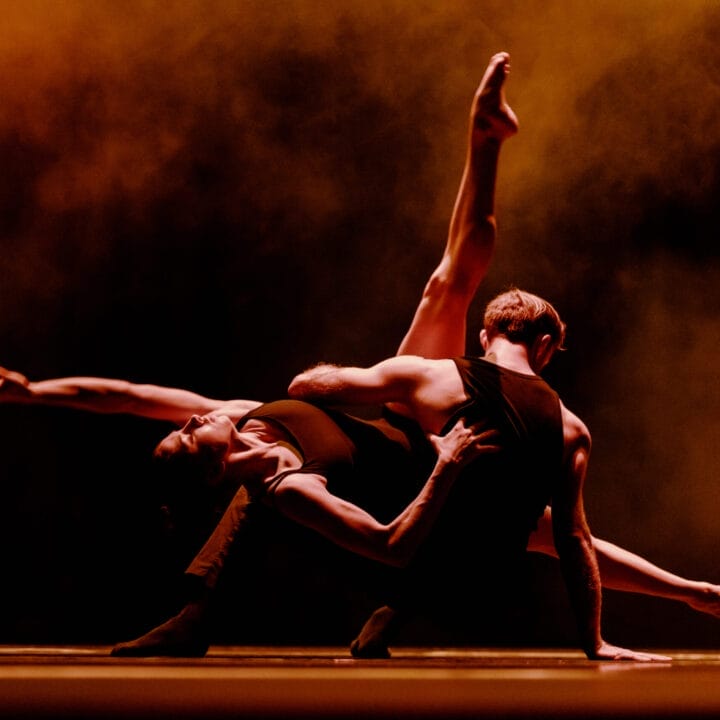 Two dancers perform a dramatic pose on stage, one arching backward with an extended leg, against a dark background with orange lighting and smoke effects.