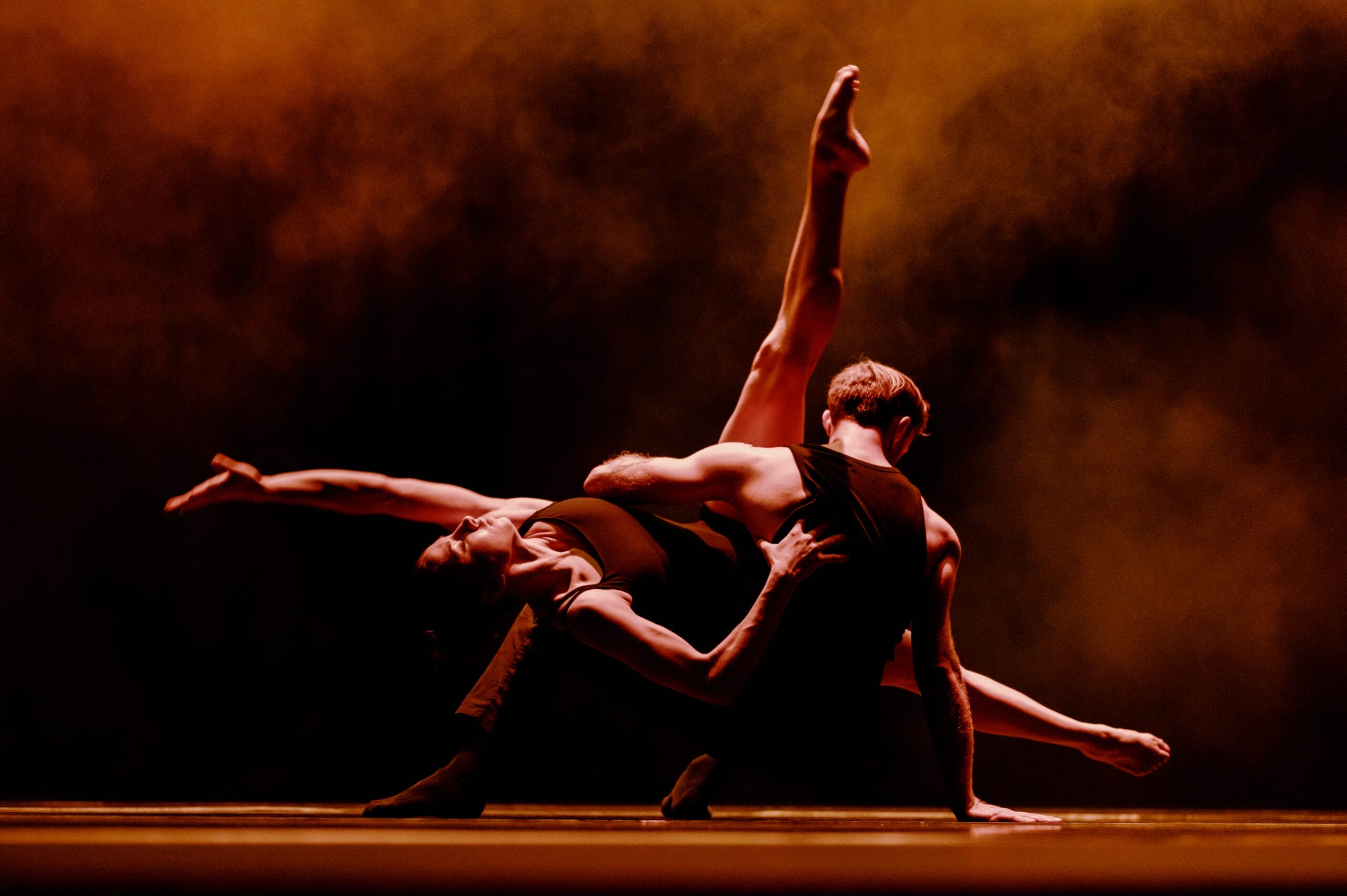 Two dancers perform a dramatic pose on stage, one arching backward with an extended leg, against a dark background with orange lighting and smoke effects.
