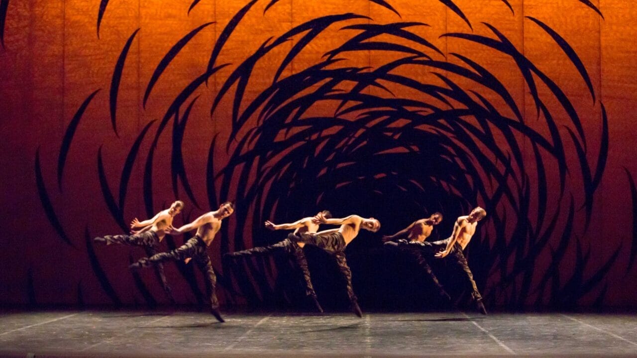 Five dancers leap mid-air on stage with an abstract orange and black swirling backdrop behind them.