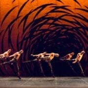 Five dancers leap mid-air on stage with an abstract orange and black swirling backdrop behind them.