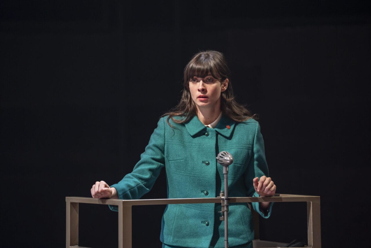 A woman in a green jacket stands behind a podium, gripping the sides and speaking into a vintage microphone, evoking the tense atmosphere of a John le Carré spy novel.
