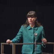 A woman in a green jacket stands behind a podium, gripping the sides and speaking into a vintage microphone, evoking the tense atmosphere of a John le Carré spy novel.